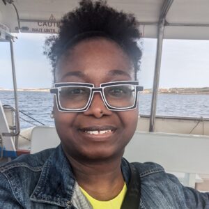 A black woman with glasses wearing a yellow top and denim jacket smiling