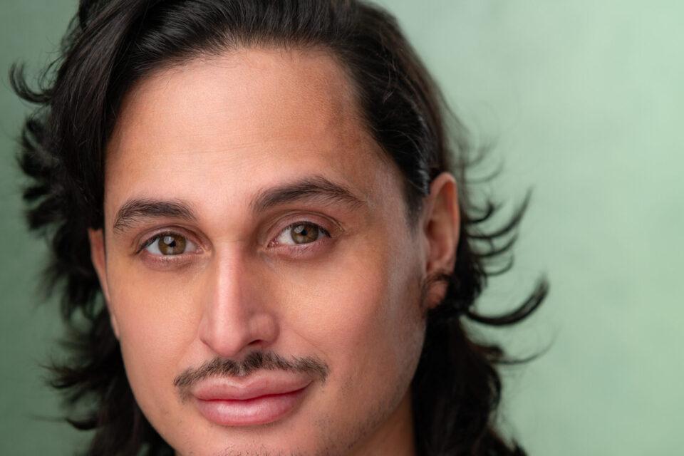 A man with long dark hair and a light brown ribbed jumper