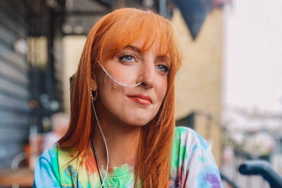 A young woman with red hair and a blue top leaning on a coffee table. She has an oxygen tube attached to her nose.
