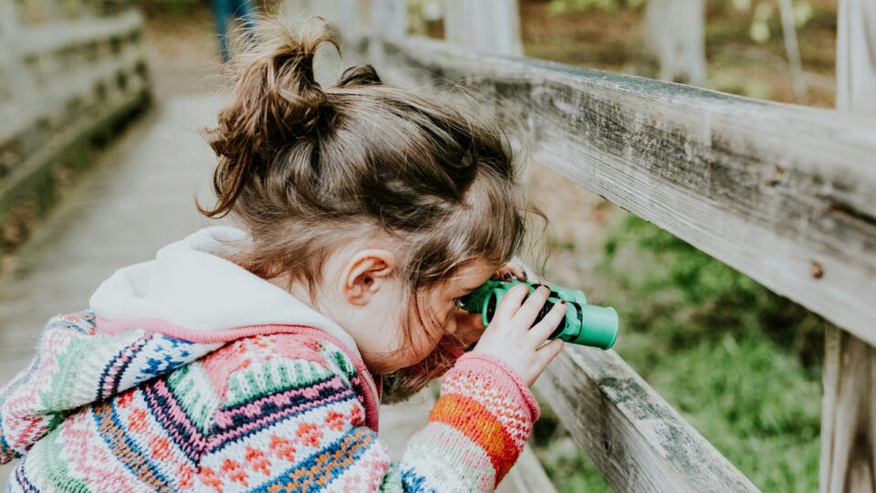 A child wearing a brightly coloured jumper is looking through green binoculars at something offscreen