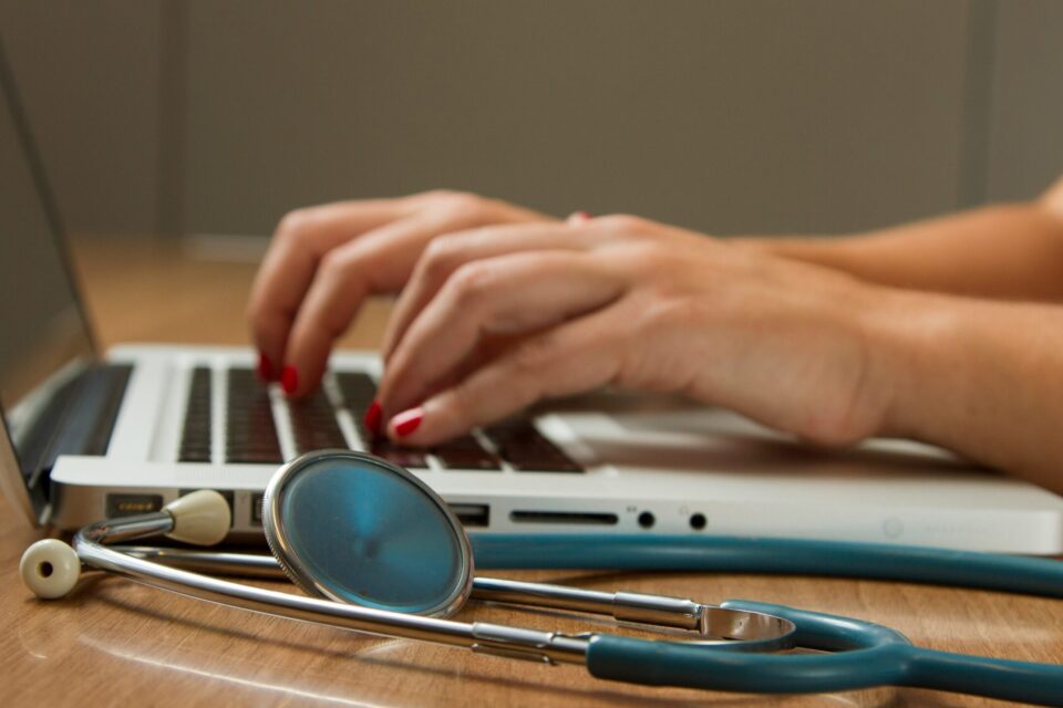 close up of hands typing on a laptop, with a stethoscope lying on the table beside it