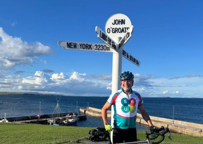 A man wearing an SRUK vest standing by the John O'Groats sign with his bike